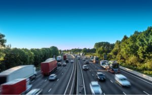 Multi-lane highway with cars and trucks; clear blue sky; green trees along roadside.
