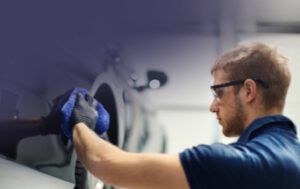 Technician polishing car headlight with blue microfiber cloth, wearing safety glasses.